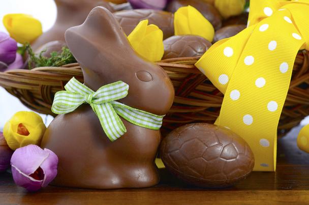 Happy Easter chocolate hamper of eggs and bunny rabbits in large basket with yellow and pink purple silk tulip flowers on dark wood table, closeup.