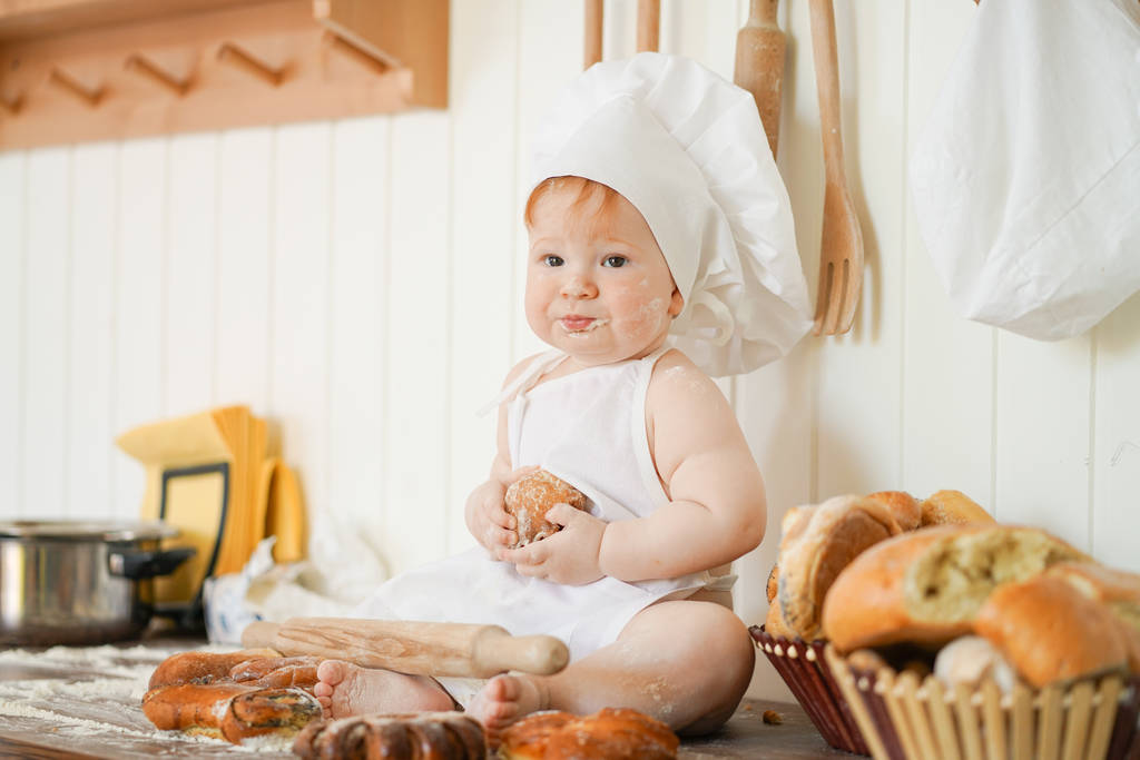 little baker child in chef hat at kitchen table alone with bread and buns